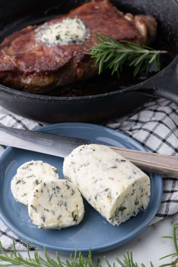 Sliced compound butter on a blue plate. A cooked steak with melted steak butter is behind the plate in a cast iron pan.