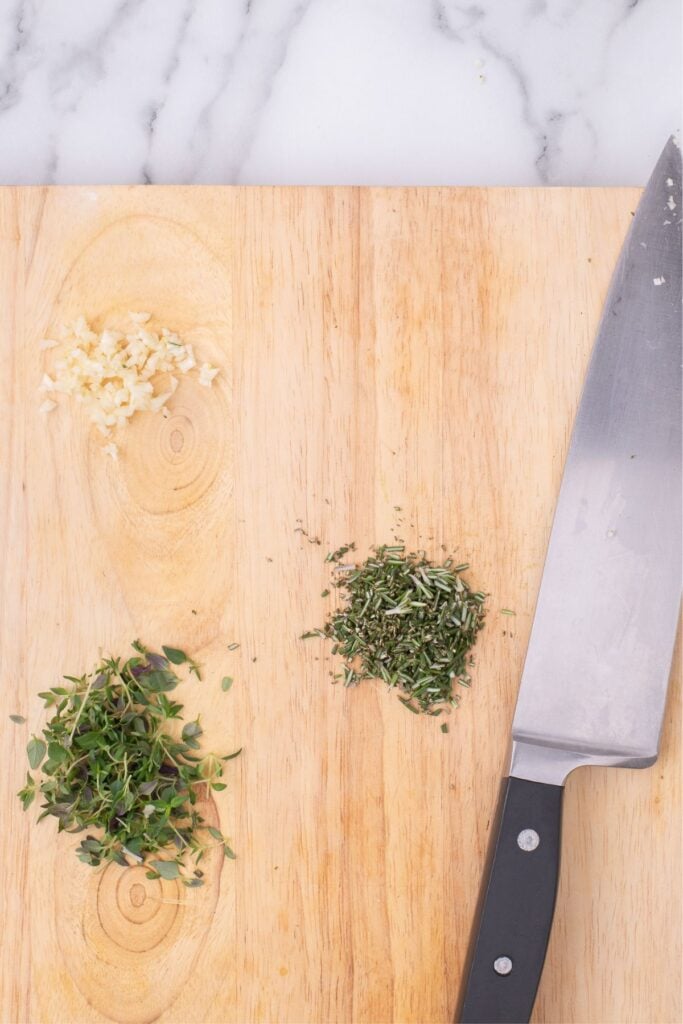 Chopped thyme, rosemary, and garlic on a wooden cutting board.