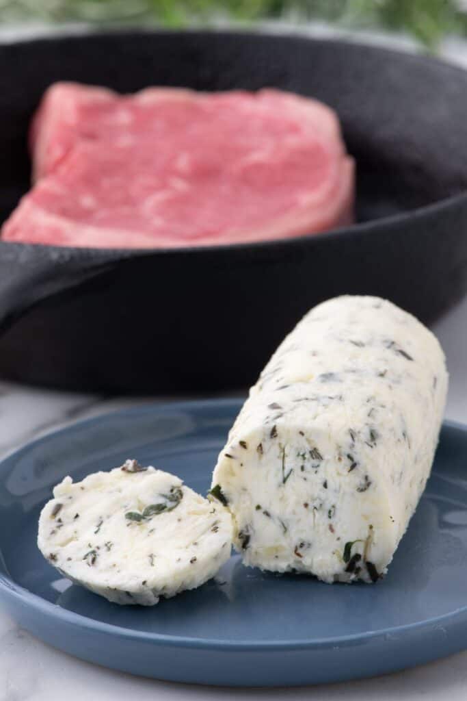 Sliced compound butter on a blue plate. A raw steak is behind the plate in a cast iron pan.