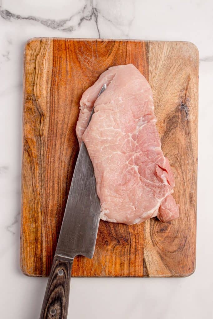 Pork chop on a wooden cutting board being sliced in half lengthwise to create a pocket for stuffing.