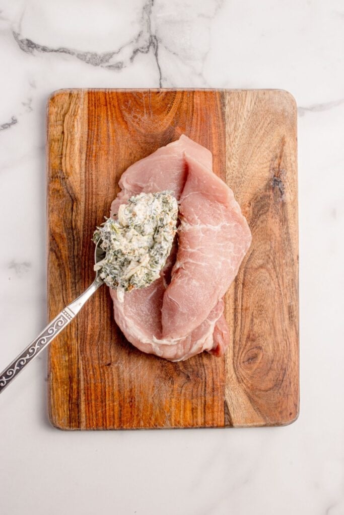 Butterflied pork chop on a wooden cutting board. A large spoonful of spinach and cheese mixture is being spooned into the center.