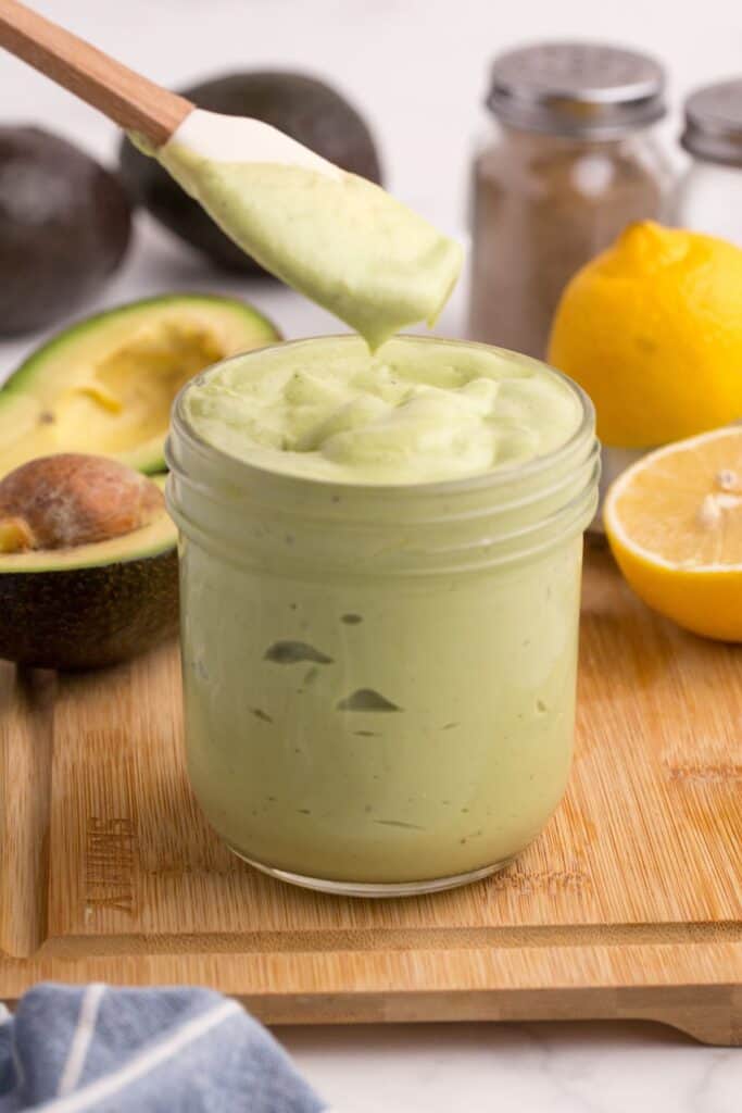 Jar of avocado mayo on a wooden cutting board. A white spatula is lifting a small amount out of the jar to show texture.