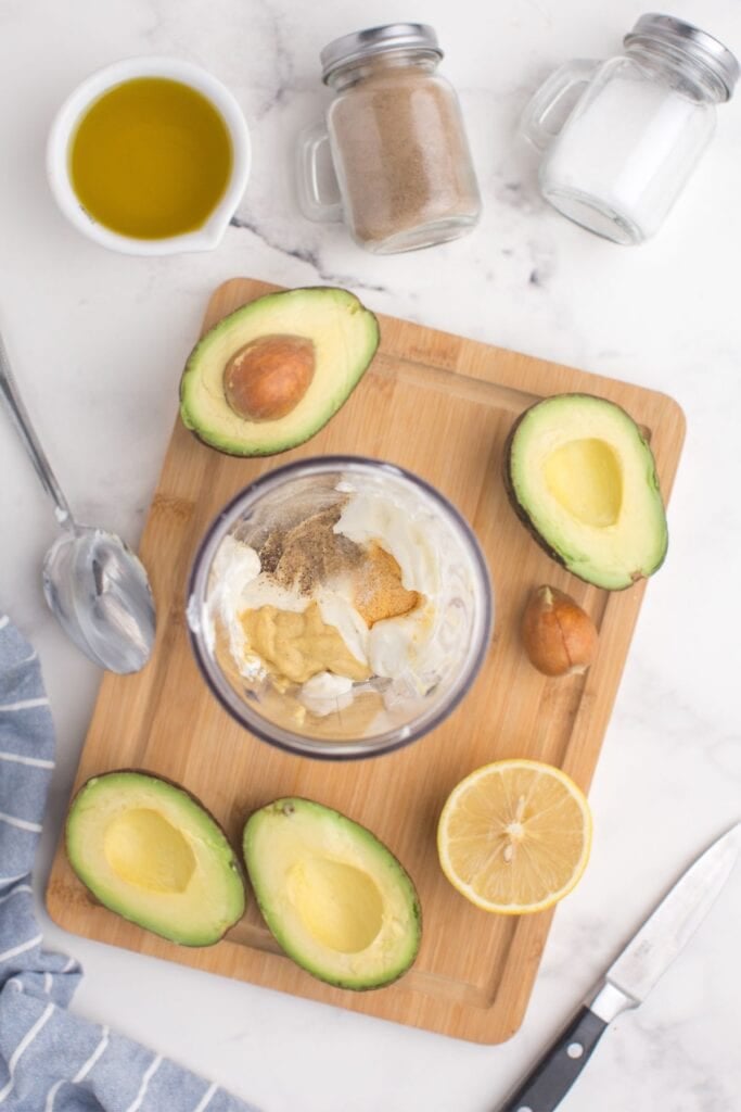 Glass jar with sour cream, lemon juice, Dijon mustard, garlic powder, salt, and pepper. Four avocado halves are surrounding the jar.