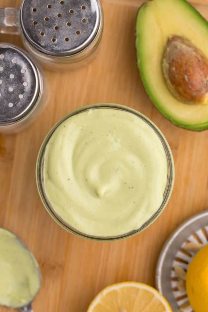 Glass jar of avocado mayo. Jar is surrounded by an avocado half, a lemon squeezer and half a lemon, and a pair of salt and pepper shakers.