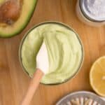 Glass jar of avocado mayo with a small white spatula in it. Jar is surrounded by an avocado half, a lemon squeezer and half a lemon, and a pair of salt and pepper shakers.