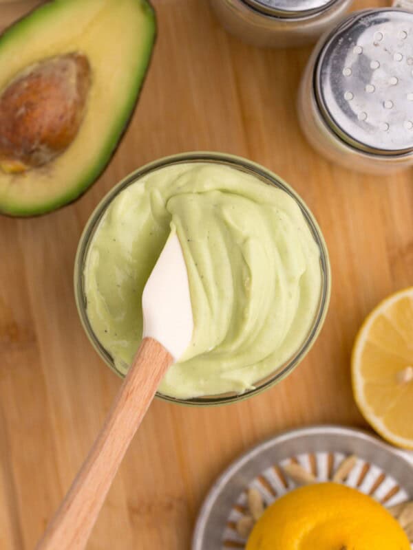 Glass jar of avocado mayo with a small white spatula in it. Jar is surrounded by an avocado half, a lemon squeezer and half a lemon, and a pair of salt and pepper shakers.