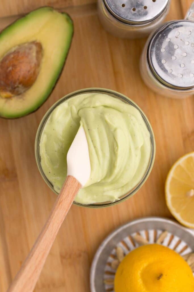 Glass jar of avocado mayo with a small white spatula in it. Jar is surrounded by an avocado half, a lemon squeezer and half a lemon, and a pair of salt and pepper shakers.