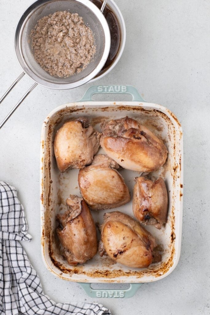 Chicken thighs have been drained of liquid and placed back in the baking dish. A bowl of marinade is next to the dish with a sieve over the top of it.