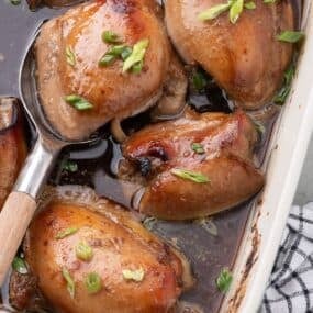 A blue baking dish with baked chicken thighs. A serving spoon is lifting a thigh out of the dish.