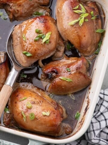 A blue baking dish with baked chicken thighs. A serving spoon is lifting a thigh out of the dish.