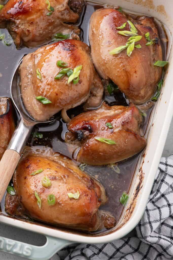 A blue baking dish with baked chicken thighs. A serving spoon is lifting a thigh out of the dish.