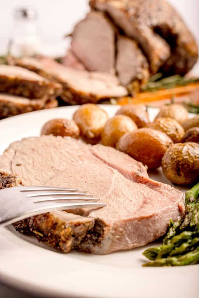 Boneless leg of lamb on a white dinner plate with baby potatoes and asparagus. A wooden cutting board with the remaining lamb is positioned behind the plate.