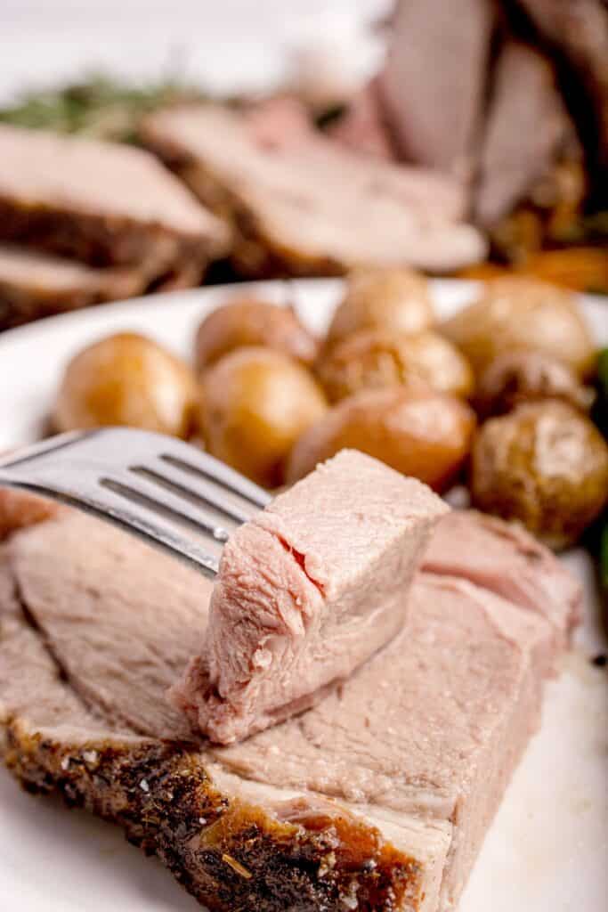 Slice of lamb on a white dinner plate with baby potatoes. A wooden cutting board with the remaining sliced leg of lamb is behind the plate.