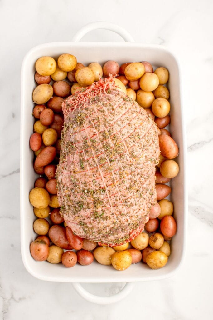 Raw, seasoned leg of lamb over a bed of raw baby potatoes.
