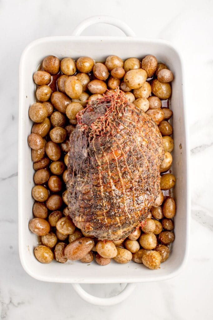 Cooked leg of lamb over a bed of baby potatoes in a large, white baking dish.