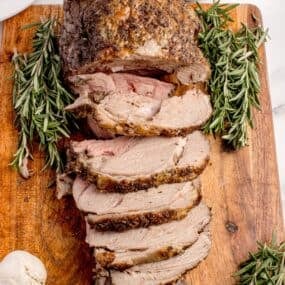 Sliced leg of lamb on a wooden cutting board, garnished with rosemary.