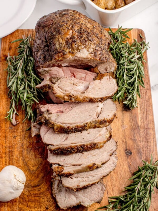 Sliced leg of lamb on a wooden cutting board, garnished with rosemary.