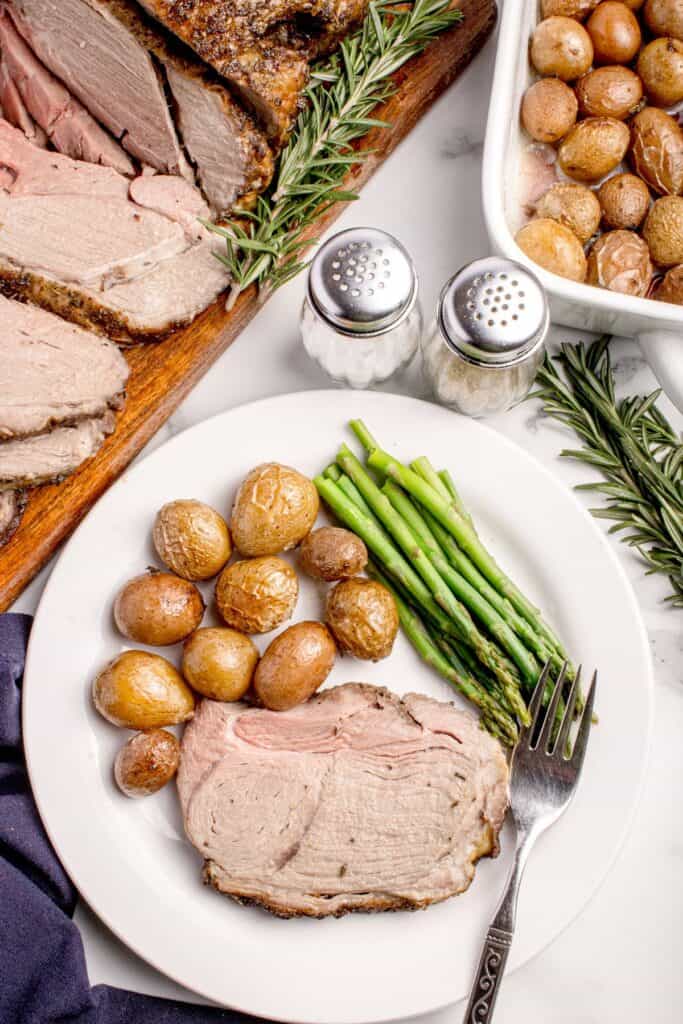 White dinner plate with a slice of lamb, baby potatoes, and asparagus. The plate is surrounded by a wooden cutting board with the remaining leg of lamb, a white baking dish with baby potatoes, and a set of salt and pepper shakers.