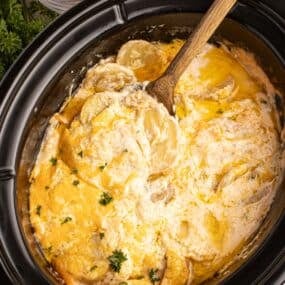 A wooden spoon scooping a serving of scalloped potatoes out of the crockpot insert.