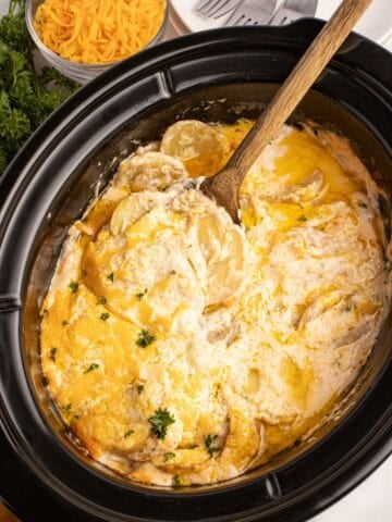 A wooden spoon scooping a serving of scalloped potatoes out of the crockpot insert.