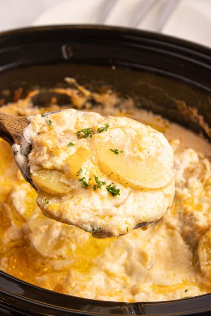 A wooden spoon lifting a serving of scalloped potatoes out of a crockpot insert.