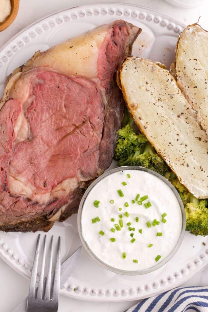 A small glass bowl of horseradish is on a white dinner plate along with a large slice of prime rib, a serving of broccoli, and a baked potato.