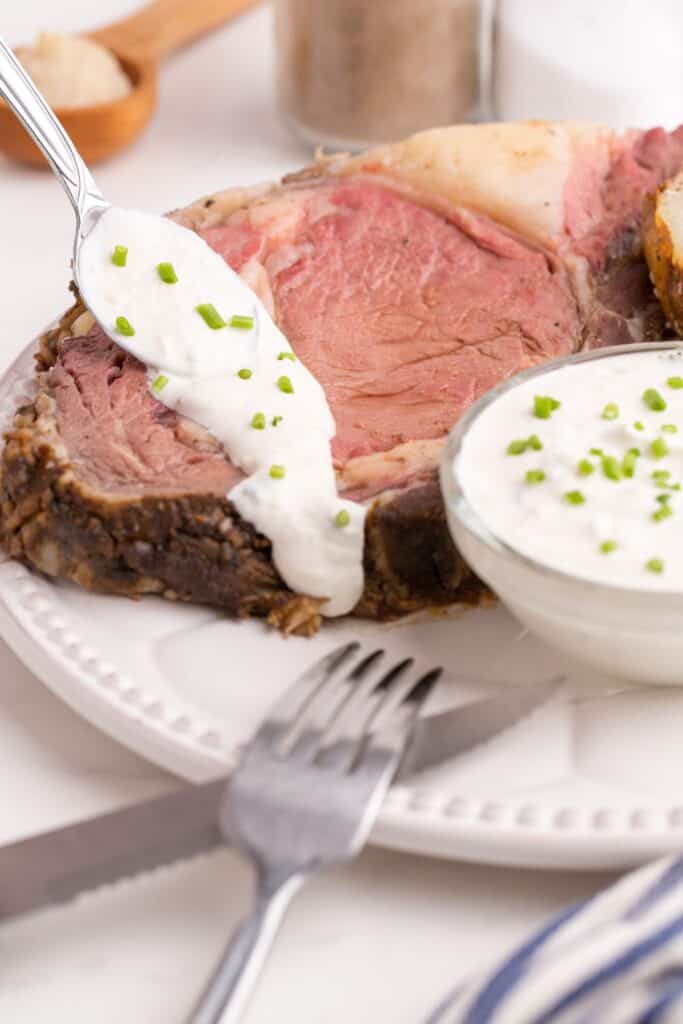 Small glass bowl of horseradish sauce on a white dinner plate with a large slice of prime rib. A spoon is pouring the horseradish over the top of the beef.