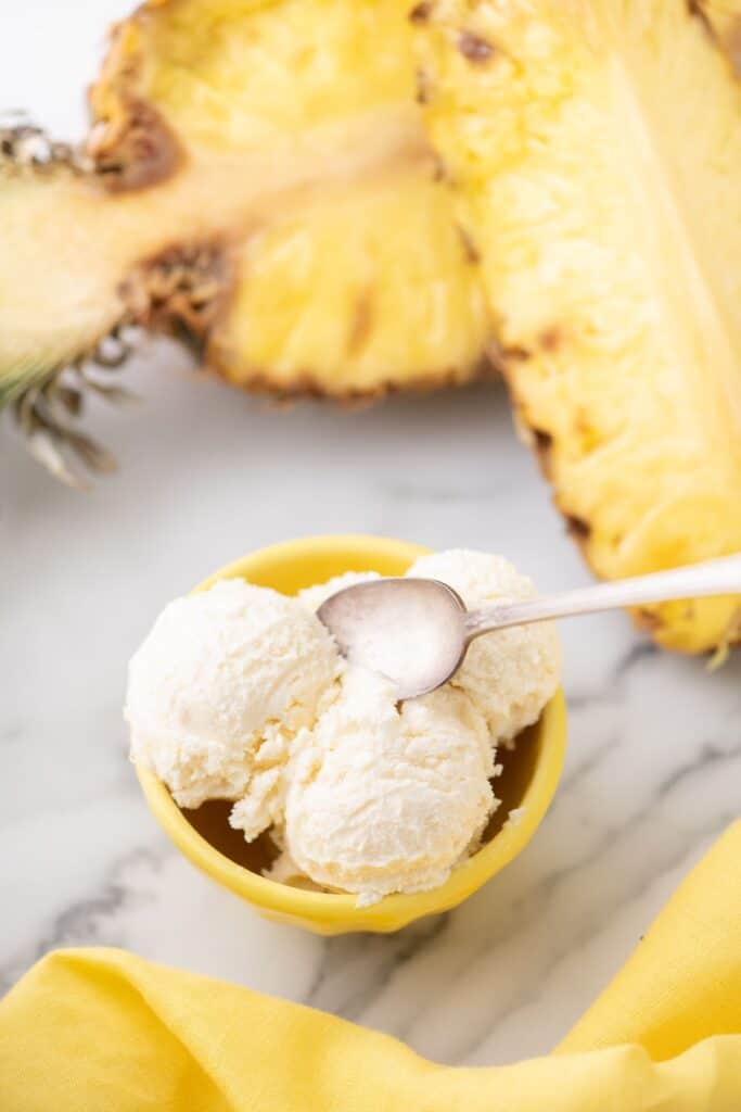 Three scoops of pineapple ice cream in a yellow bowl with a spoon. Bowl is surrounded by two halves of a pineapple and a yellow tea towel.