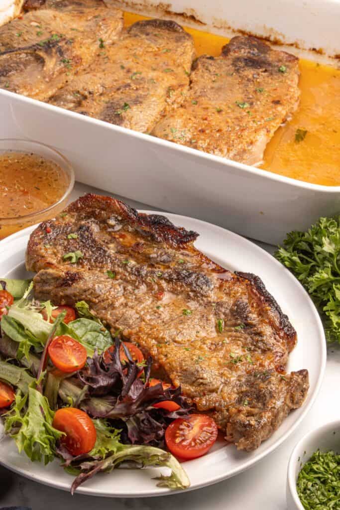 A white dinner plate with pork steaks and a salad. Behind the plate is a large white baking dish with the remaining cooked pork steaks in it.
