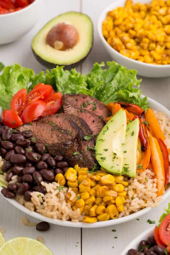 Completed steak bowl with sliced flank steak, bell peppers, black beans, cherry tomatoes, corn, and sliced avocado over a bed of rice.
