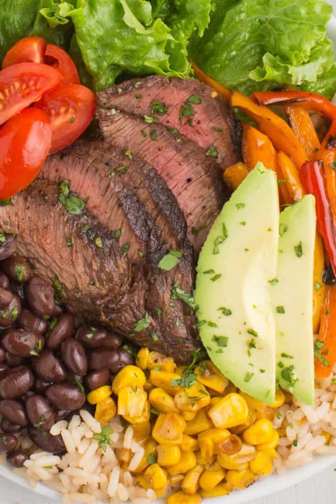 Completed steak bowl with sliced flank steak, bell peppers, black beans, cherry tomatoes, corn, and sliced avocado over a bed of rice.