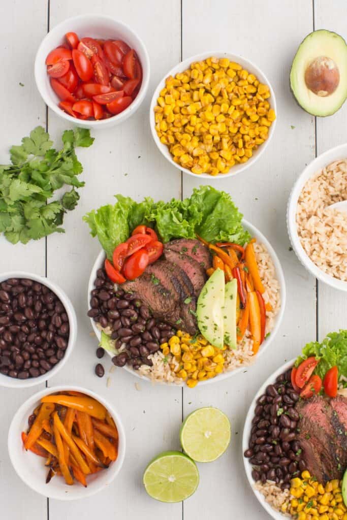 Two steak bowls surrounded by white serving bowls of rice, corn, tomatoes, beans, and bell peppers. Sliced avocado, limes, and cilantro are placed among the bowls.
