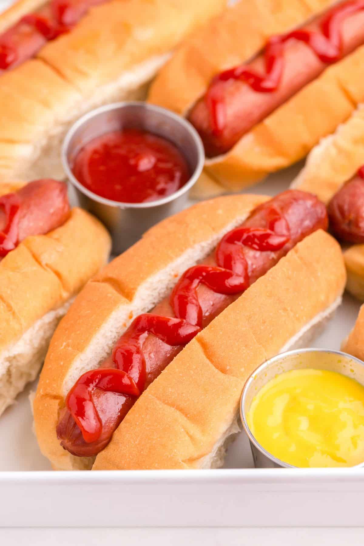 Several hot dogs are lined up on a platter with little cups of ketchup and mustard.