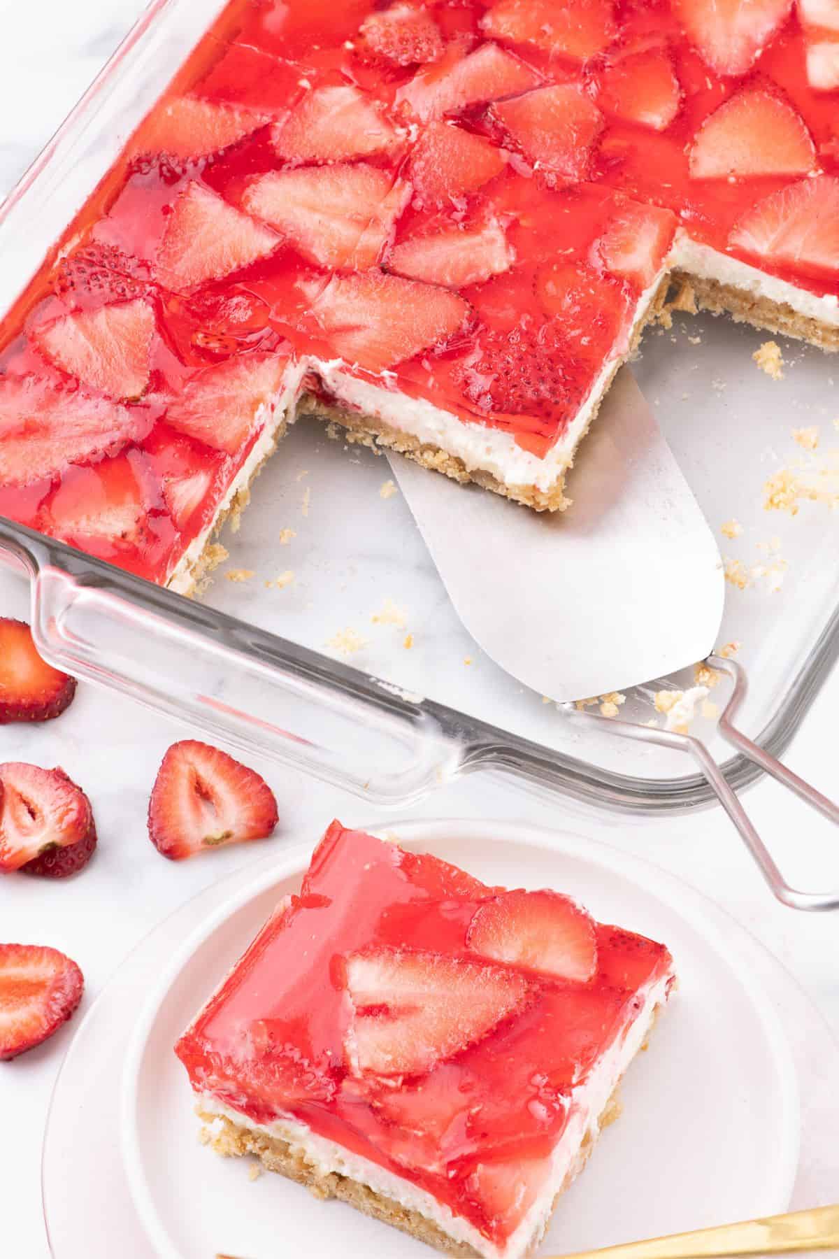 Strawberry pretzel salad sliced and served from a glass baking dish. A white plate in front of the baking dish has a piece of the dessert on it.