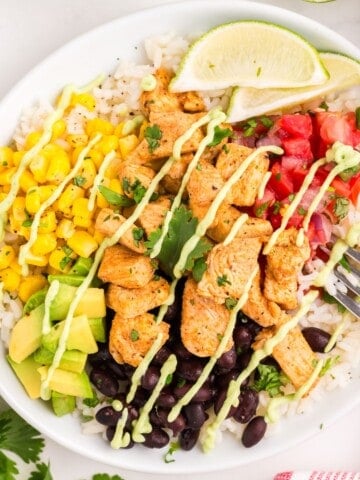 Finished burrito bowl in a white bowl with rice, seasoned chicken, black beans, corn, avocado, pico de gallo, lime wedges and avocado sauce.