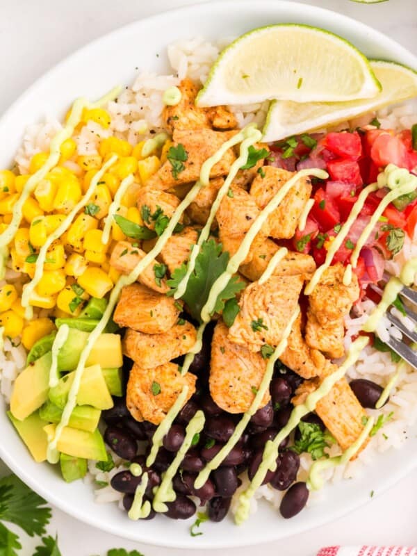 Finished burrito bowl in a white bowl with rice, seasoned chicken, black beans, corn, avocado, pico de gallo, lime wedges and avocado sauce.