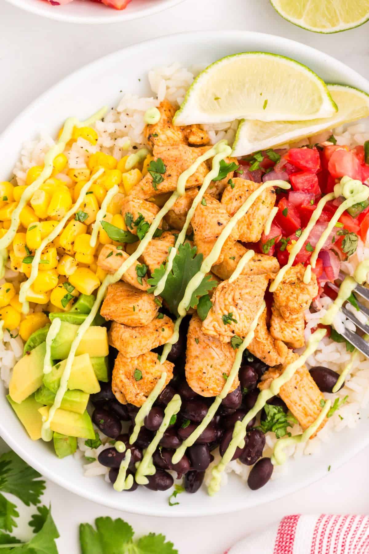 Finished burrito bowl in a white bowl with rice, seasoned chicken, black beans, corn, avocado, pico de gallo, lime wedges and avocado sauce.