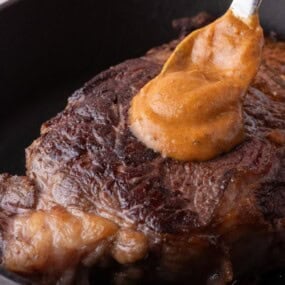 Cooked steak in a cast iron pan. A spoon is dolloping a serving of cowboy butter on top.