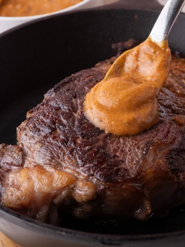 Cooked steak in a cast iron pan. A spoon is dolloping a serving of cowboy butter on top.
