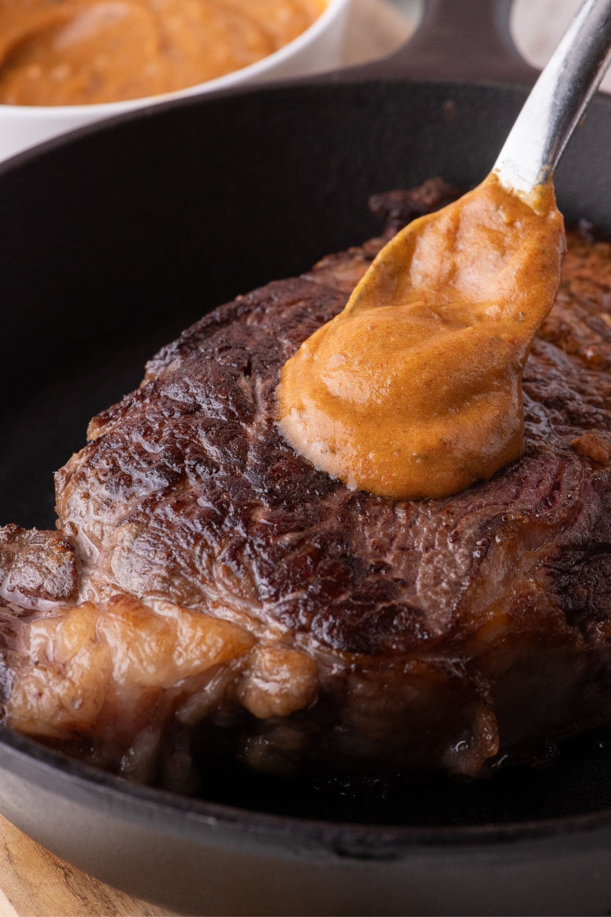 Cooked steak in a cast iron pan. A spoon is dolloping a serving of cowboy butter on top.