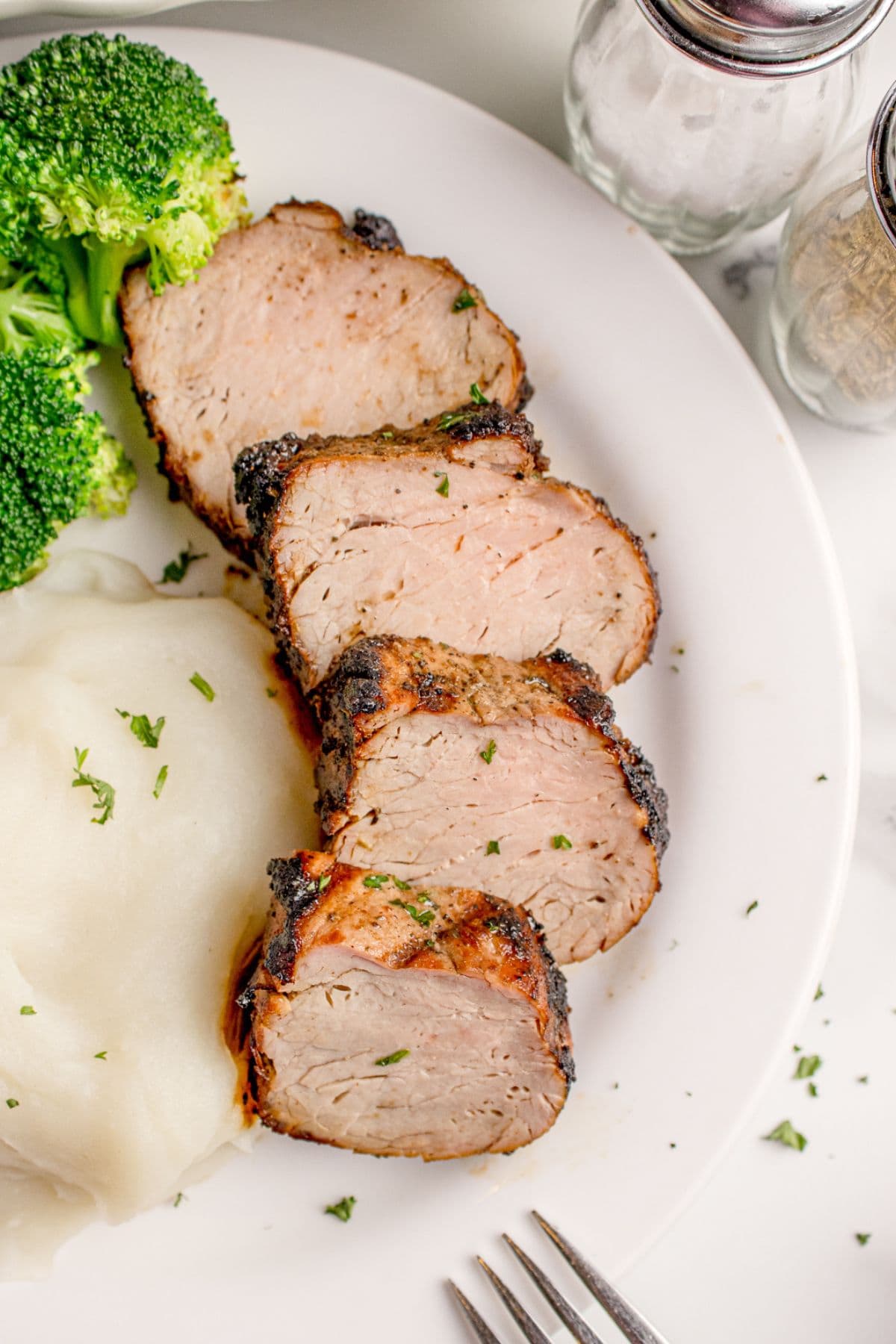 Sliced grilled pork tenderloin on a white dinner plate with a side of mashed potatoes and broccoli.