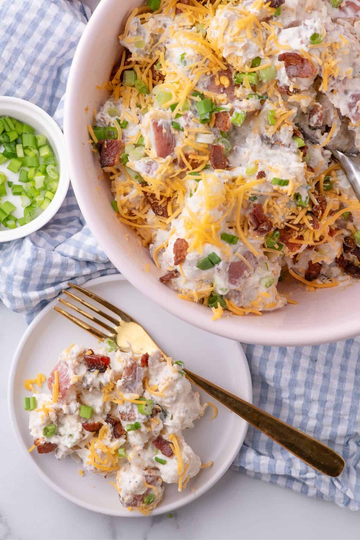 A white dinner plate with loaded potato salad on it. A serving bowl is next to the plate with the rest of the potato salad.