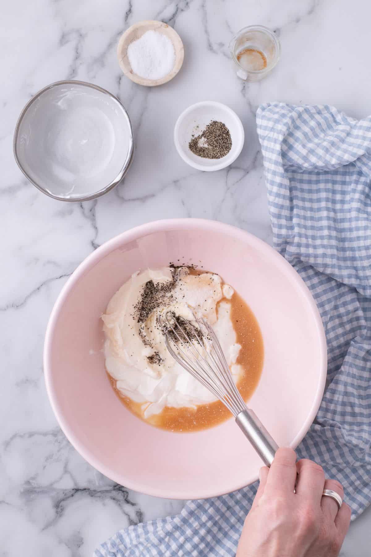 A large mixing bowl with sour cream, mayonnaise, apple cider vinegar, salt, and pepper being whisked together.