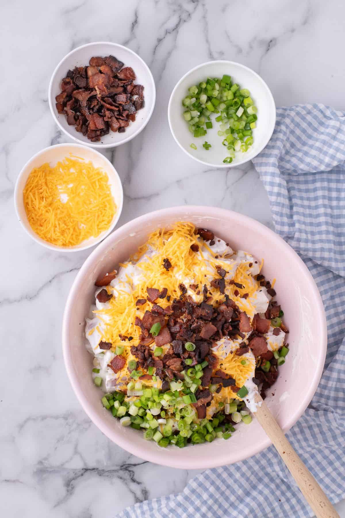 Large mixing bowl with diced potatoes, sour cream mixture, and topped with shredded cheese, bacon bits, and chopped green onions.