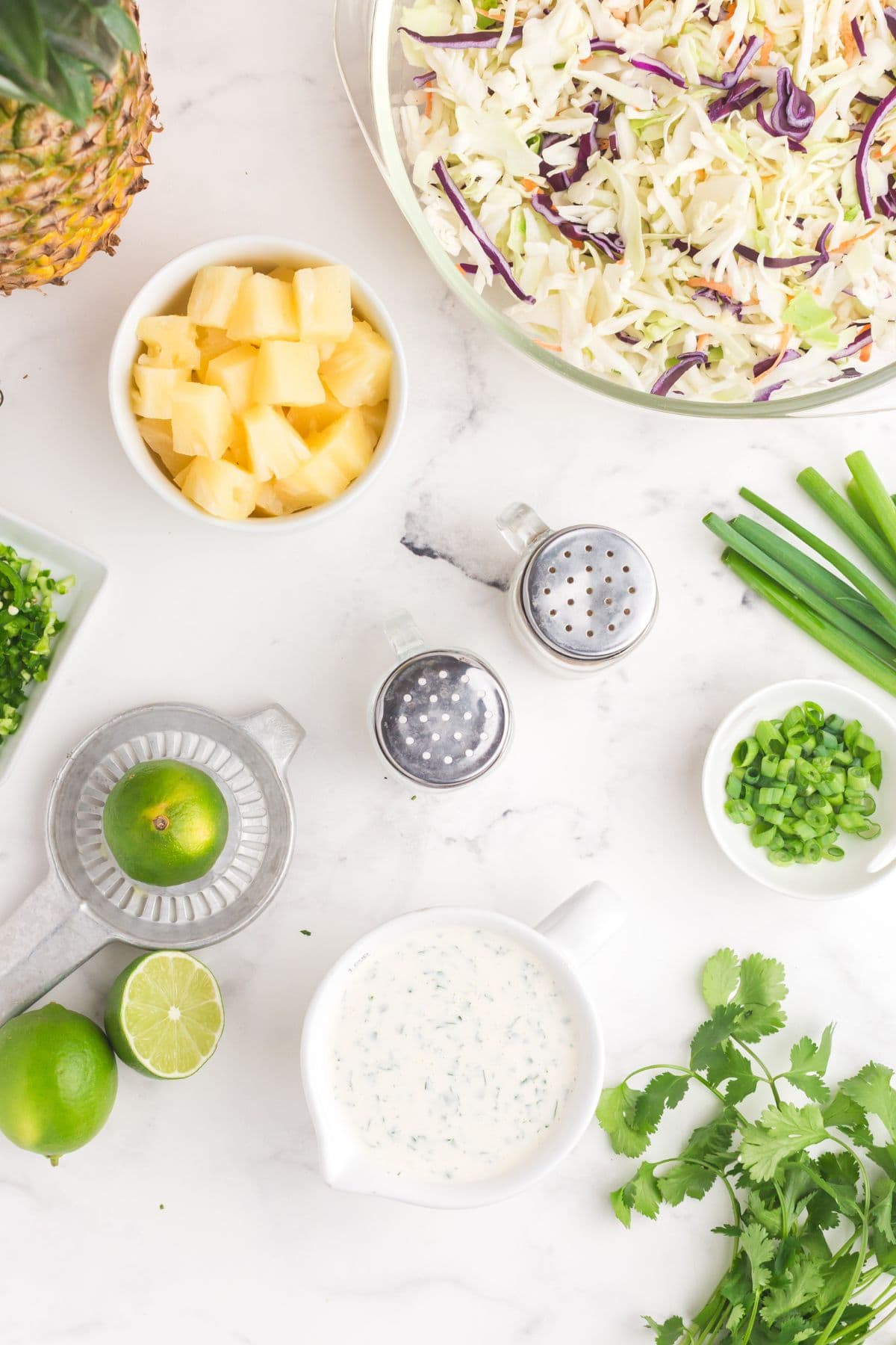 White pitcher with coleslaw dressing surrounded by cilantro sprigs, limes, chopped green onion, chopped jalapenos, a small bowl of pineapple tidbits, and a glass bowl of coleslaw mix.