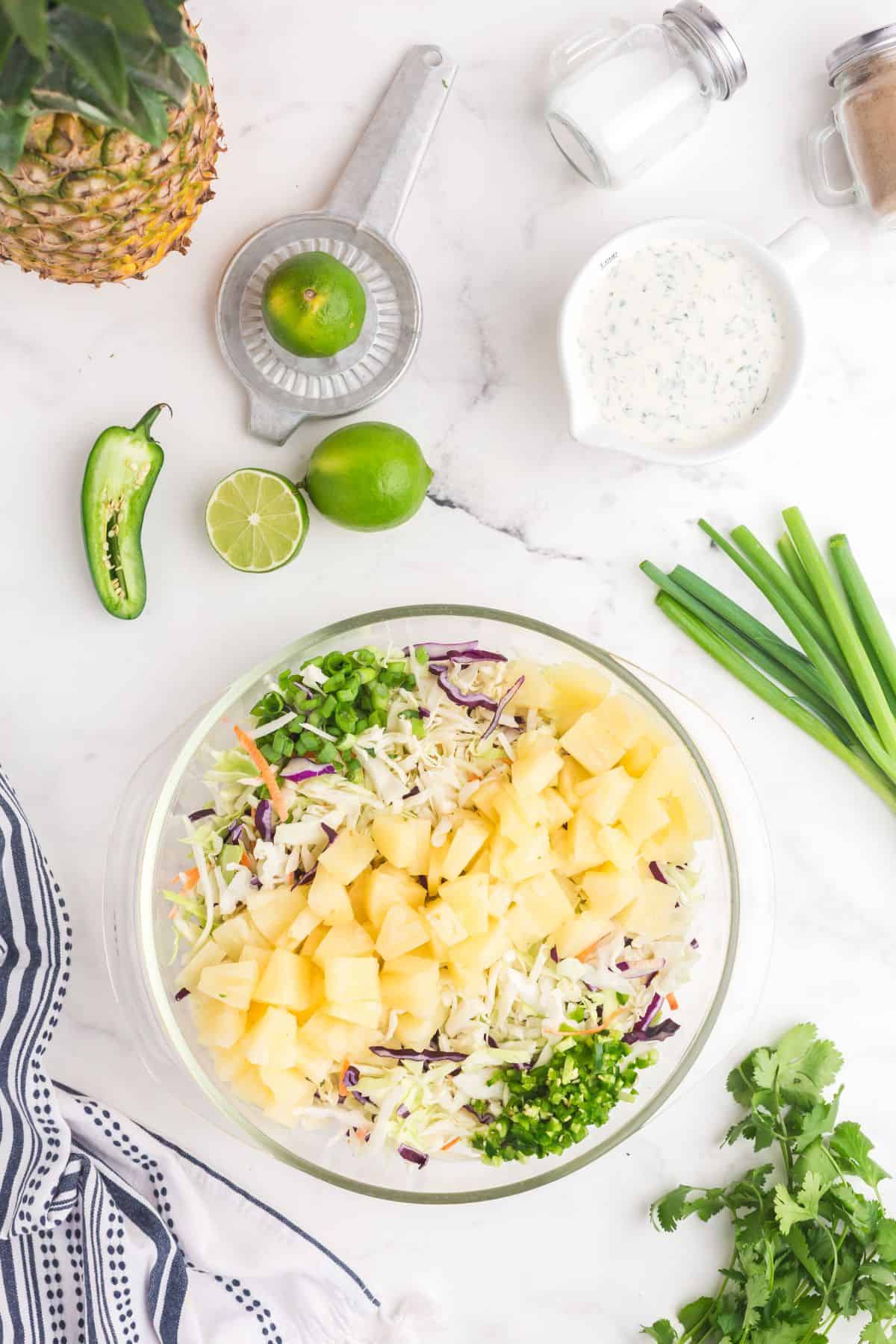 Large glass bowl of coleslaw mix with layers of chopped jalapenos, pineapple tidbits, and chopped green onions on top.