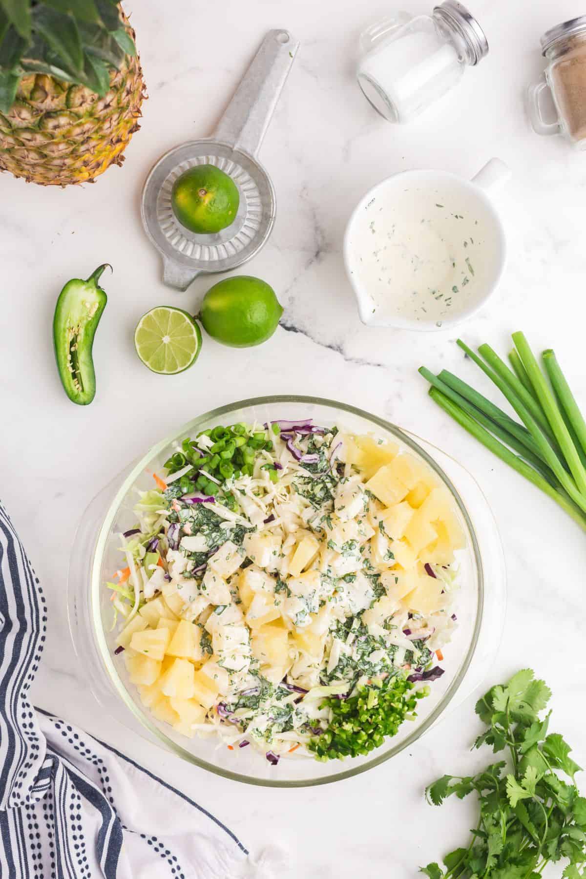 Glass mixing bowl with coleslaw mix, chopped jalapenos, chopped green onions, pineapple tidbits, and the homemade pineapple dressing.