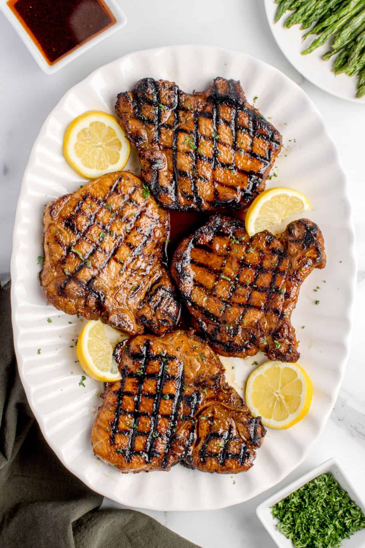 Four completed pork chops on a white serving platter, garnished with lemon slices and parsley.