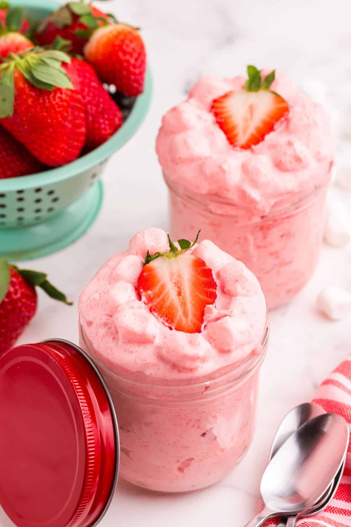 Two servings of strawberry fluff in small glass jars. A colander of strawberries sits behind the jars.