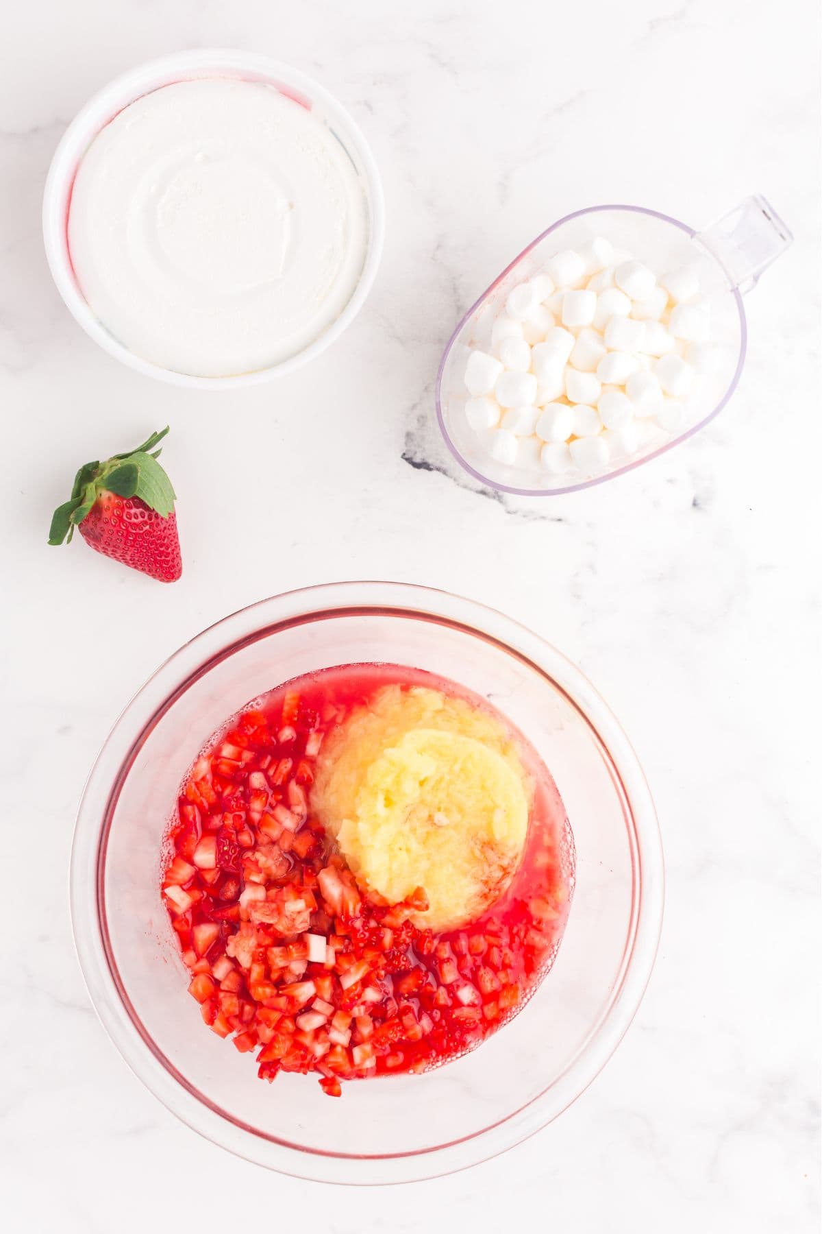Glass mixing bowl with strawberry jello, pineapple juice, chopped strawberries, and crushed pineapple.